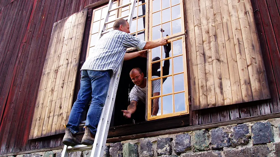 Två personer renoverar äldre träfönster på röd byggnad 