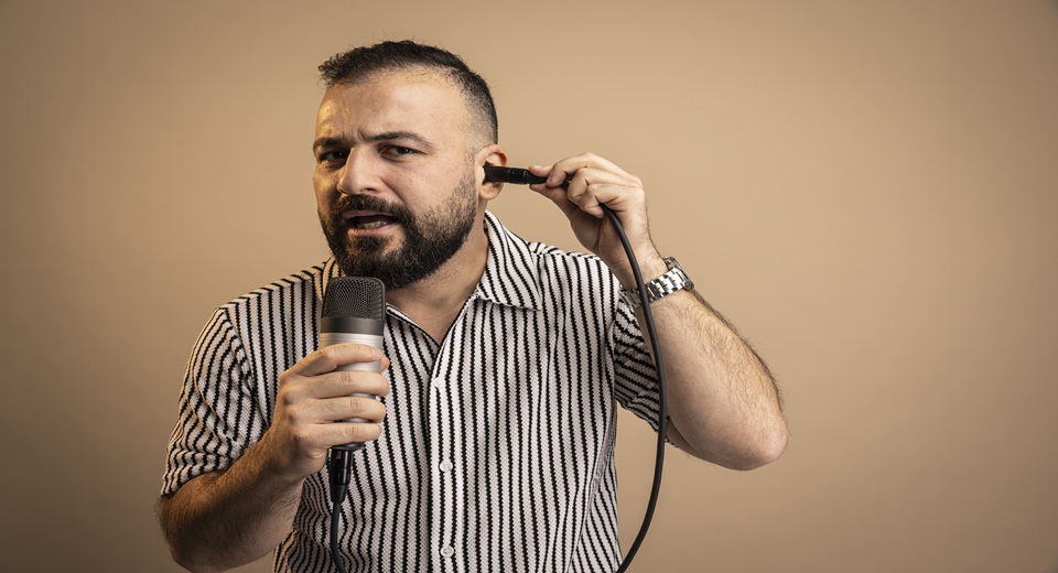 Diyari Mahmoud tittar in i kameran. Håller en mikrofon i ena handen. Håller mikrofonkontakten mot ena örat.