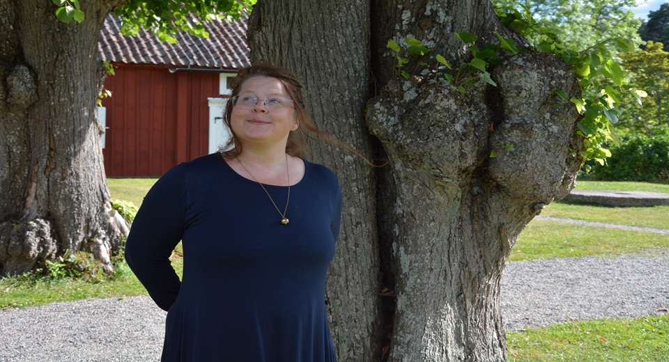 Tora Wall står leende med händerna bakom ryggen, framför ett stort träd.