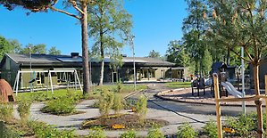 Förskolegård med träd, grönska och lekplats, förskolebyggnad i bakgrunden